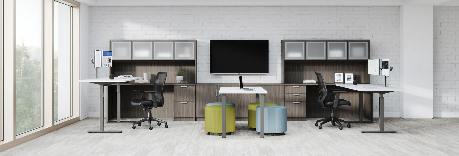 A modern office furniture setup featuring two height-adjustable standing desks with black office chairs, matching storage cabinets, a central wall-mounted monitor, and two colorful stools in yellow and blue, set against a light gray brick wall.